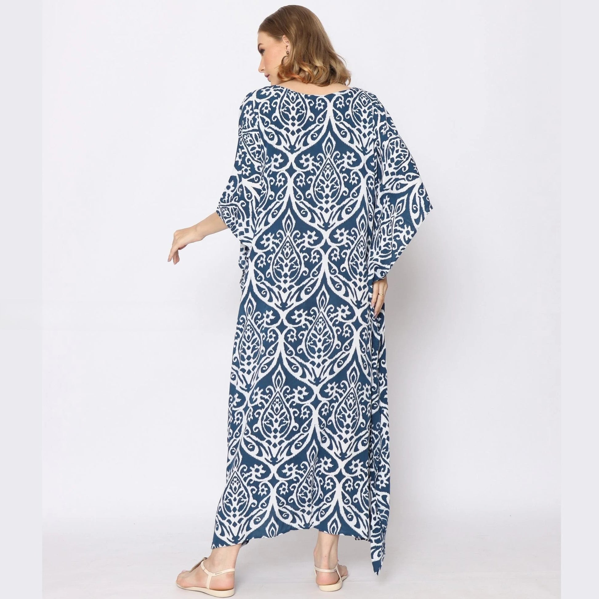 A woman is standing with her back to the camera, wearing a long, loose-fitting blue and white patterned dress. The caftan dress features an intricate white design on a dark blue background. She is wearing sandals and has shoulder-length brown hair.