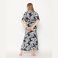 A person with long hair stands facing away, wrapped in a black and white palm leaf patterned dress. They have their hands clasped behind their back and are wearing white sandals. This stylish ensemble, akin to trendy wrap dresses for women, is set against a plain white background.