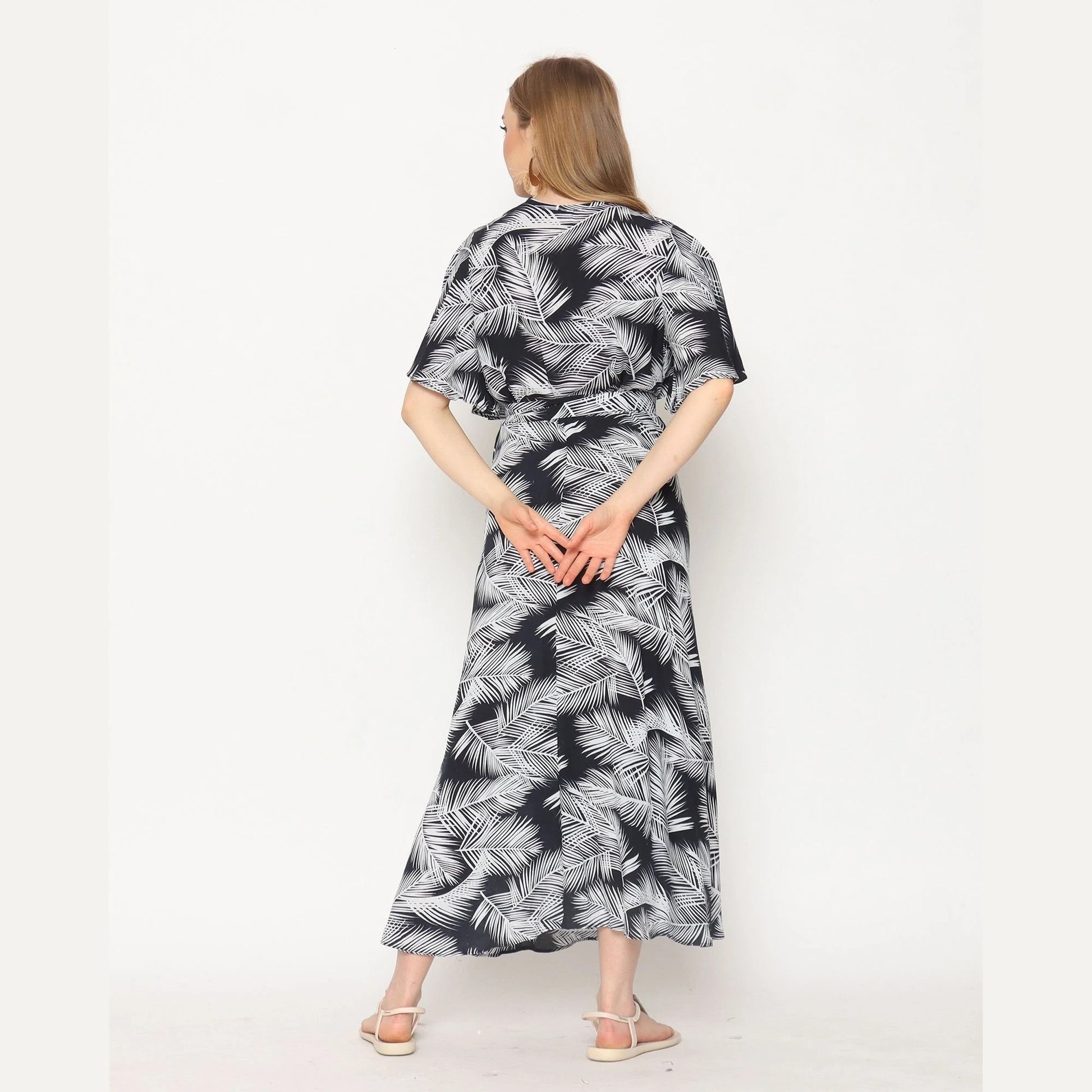 A person with long hair stands facing away, wrapped in a black and white palm leaf patterned dress. They have their hands clasped behind their back and are wearing white sandals. This stylish ensemble, akin to trendy wrap dresses for women, is set against a plain white background.