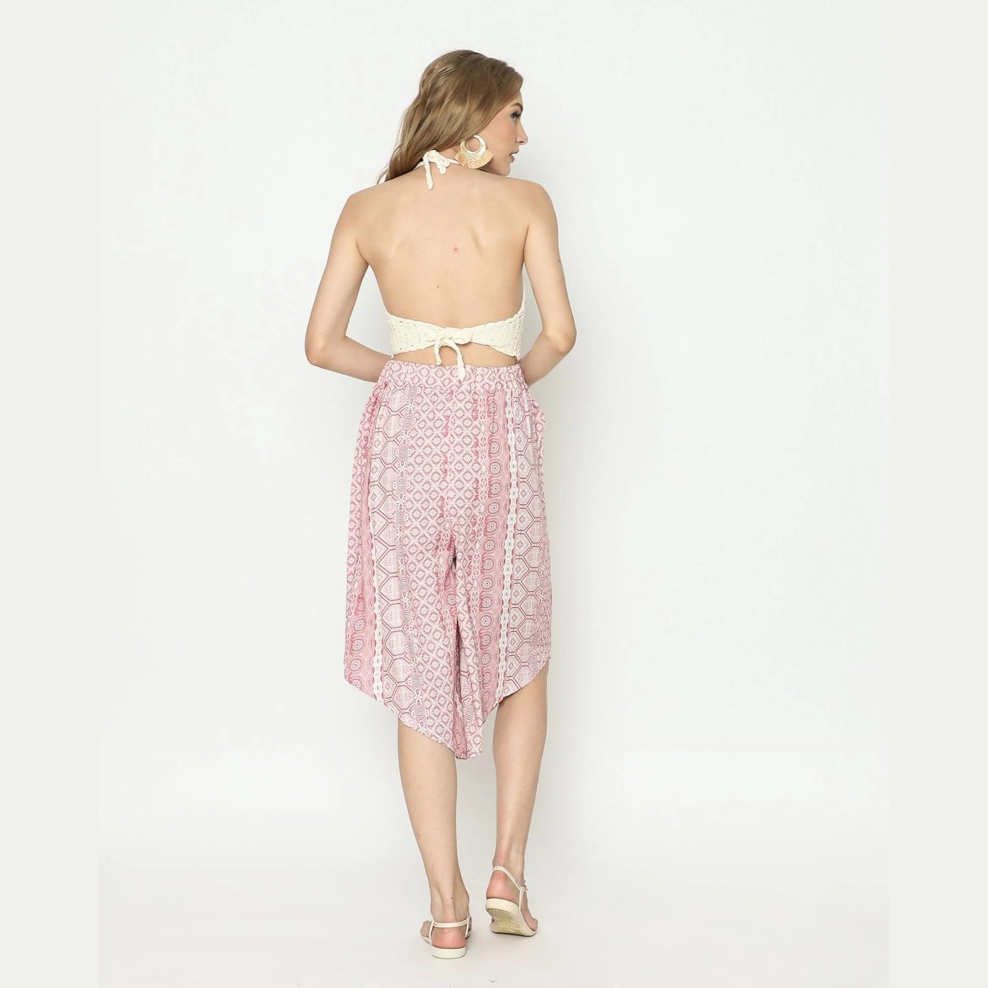 A woman stands with her back to the camera, wearing a white halter top and pink patterned skirt that elegantly contrasts with her white sandals and large earrings. Her long hair cascades down, completing the chic look against a plain white backdrop.