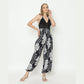 A woman in a black lace halter top and black and white floral pants poses against a plain background. She is wearing white sandals and has her left hand touching her hair and her right hand on her hip.