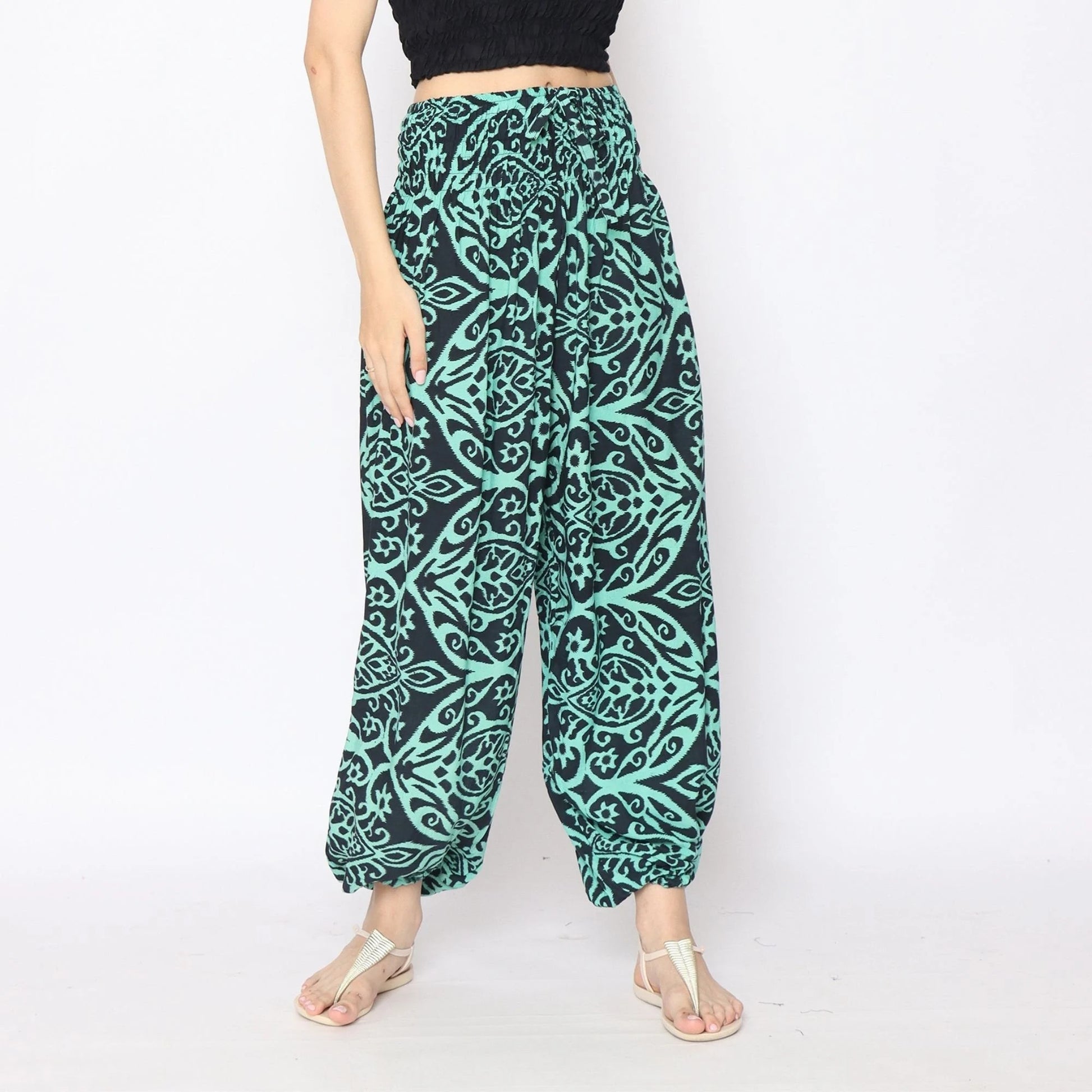 A person wearing teal and black patterned harem yoga pants and a black crop top stands against a white background. They are also wearing white sandals.