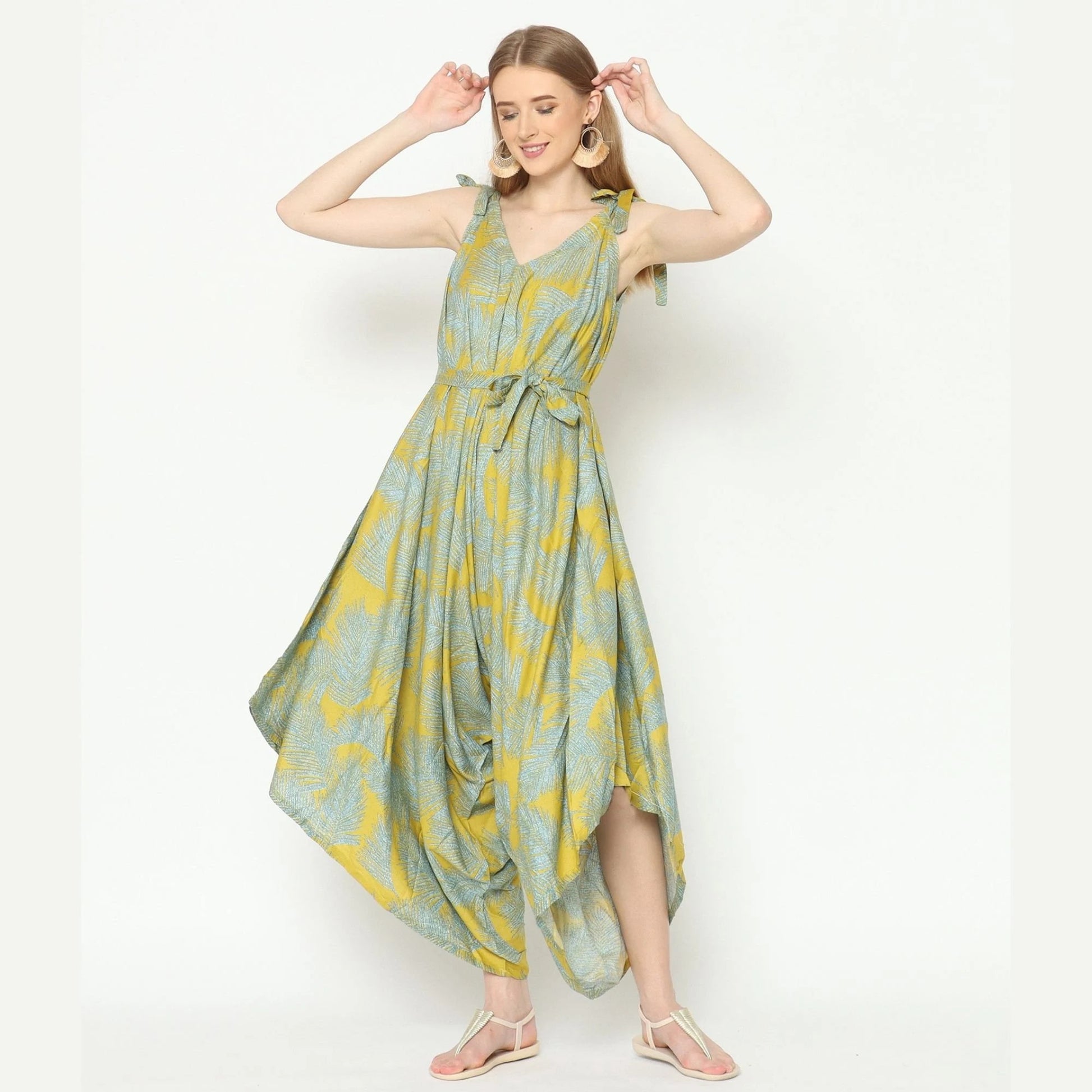 A woman in a flowing sleeveless ensemble, reminiscent of a long jumpsuit with a green and yellow pattern, poses against a plain backdrop. She wears sandals, her hands gently raised near her head, smiling softly.