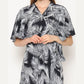 Person wearing a black and white patterned wrap dress with palm leaf designs, standing against a plain background. The dress features short, flared sleeves and a V-neckline. The individuals hands are gently clasped together.