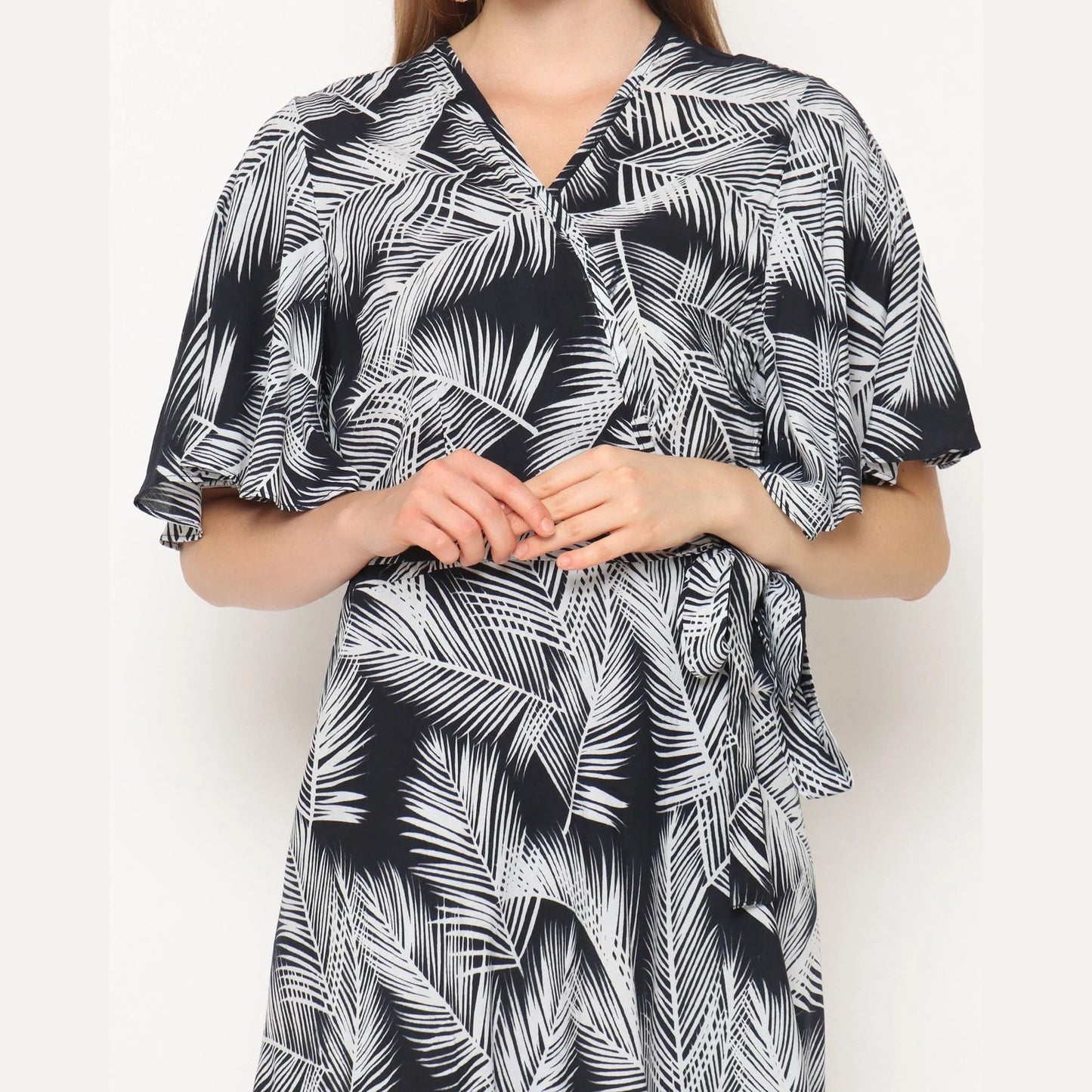 Person wearing a black and white patterned wrap dress with palm leaf designs, standing against a plain background. The dress features short, flared sleeves and a V-neckline. The individuals hands are gently clasped together.