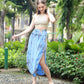 A woman in a beige crochet crop top and summer skirt with pockets strolls along a stone path in a garden. She wears white sandals, her hand gently touching her hair. Greenery and trees encircle her, with a building peeking through the lush backdrop.