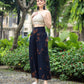 Person standing on a pebble path by green plants, wearing a beige crochet top and black boho flare pants. Theyre also in white sandals with platform soles. Background features trees and buildings.