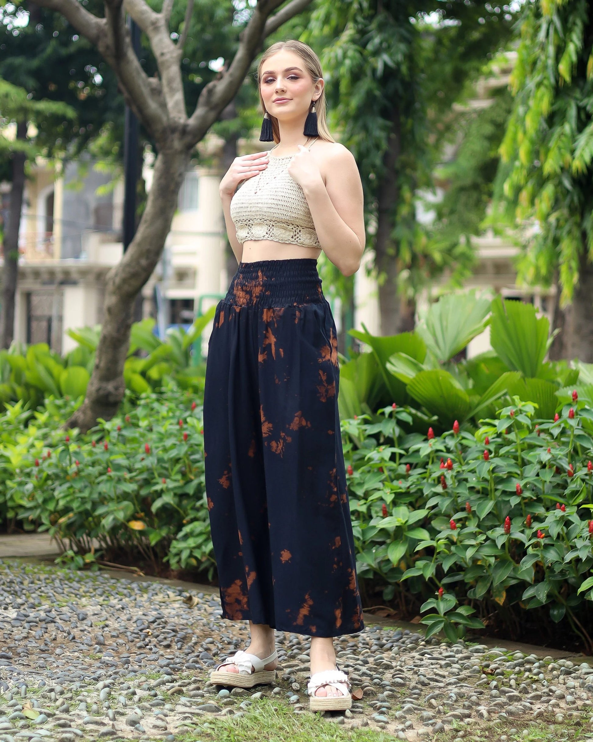 Person standing on a pebble path by green plants, wearing a beige crochet top and black boho flare pants. Theyre also in white sandals with platform soles. Background features trees and buildings.