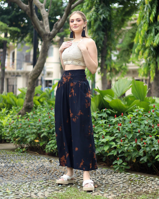 Person standing on a pebble path by green plants, wearing a beige crochet top and black boho flare pants. Theyre also in white sandals with platform soles. Background features trees and buildings.