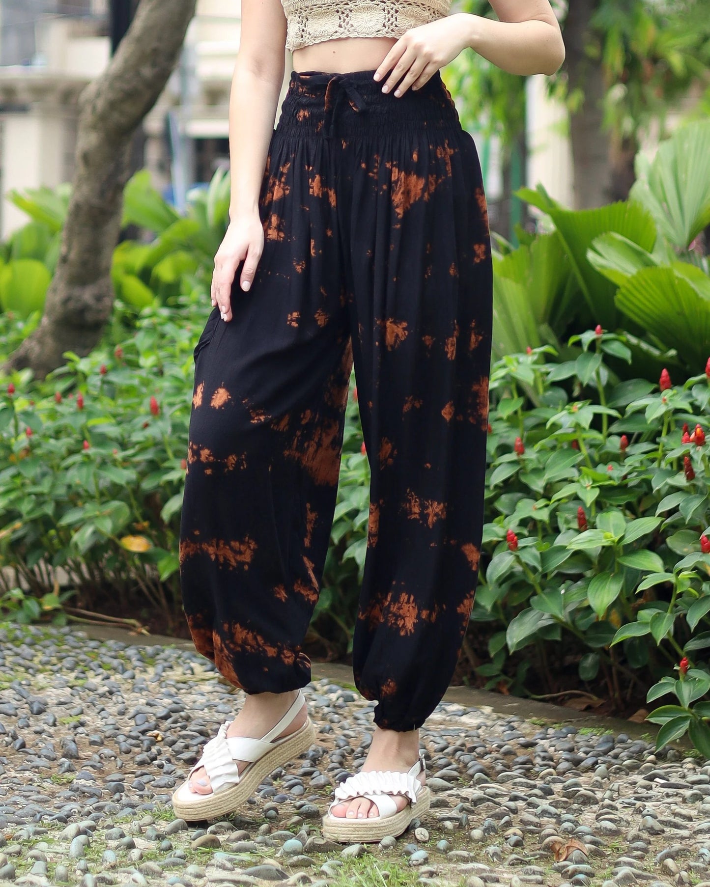 A person wearing black tie-dye yoga pants stands on a pebble path, embraced by vibrant green plants and red flowers. They are styled in a beige crochet top and white sandals, with arms relaxed by their sides.
