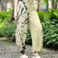 A person in light yellow tie-dye yoga pants for women and a black crop top stands on a stone pathway. They are wearing white sandals. Green foliage and small red flowers are in the background, creating a vibrant contrast to their ensemble.