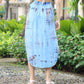 Rear view of a person wearing a light blue tie-dye dress with a bare back, akin to a summer skirt with pockets, standing on a pebble pathway. They are wearing sandals and surrounded by lush greenery, including bushes and small trees in an inviting outdoor setting.
