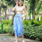A woman in a tan crochet crop top and a flowing summer skirt with pockets stands on a pebble path in a lush garden. She is looking down while touching her hair, surrounded by green plants and trees. Shes wearing white sandals.