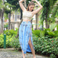A person poses outdoors, smiling with arms raised. They wear a crochet crop top and a blue patterned summer skirt with pockets, featuring a high slit, along with white sandals. The background is lush with greenery and trees.
