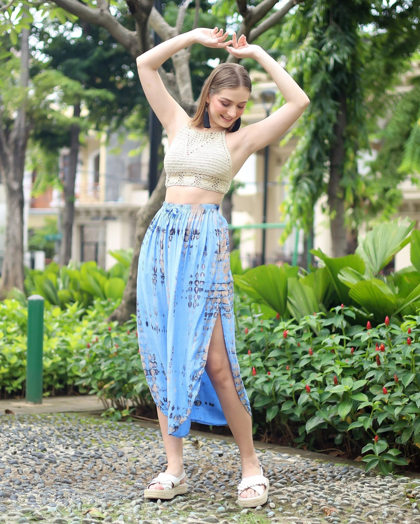 A person poses outdoors, smiling with arms raised. They wear a crochet crop top and a blue patterned summer skirt with pockets, featuring a high slit, along with white sandals. The background is lush with greenery and trees.