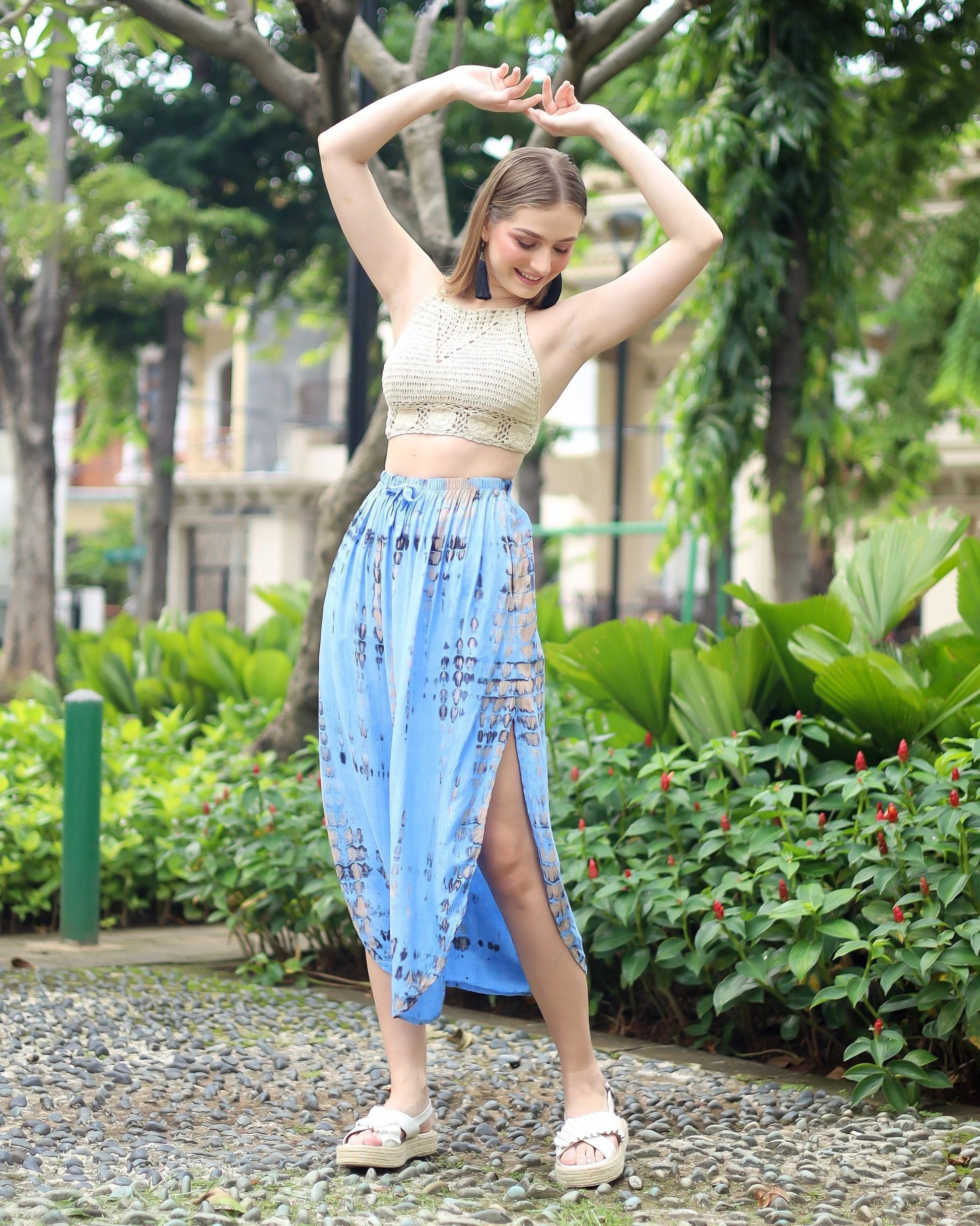 A person poses outdoors, smiling with arms raised. They wear a crochet crop top and a blue patterned summer skirt with pockets, featuring a high slit, along with white sandals. The background is lush with greenery and trees.