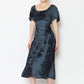 A person smiles, standing against a plain background in a striking tie-back dress. The blue-and-black tie-dye outfit features short sleeves and a square neckline. They complement the look with sandals, and their hair is neatly tied back.