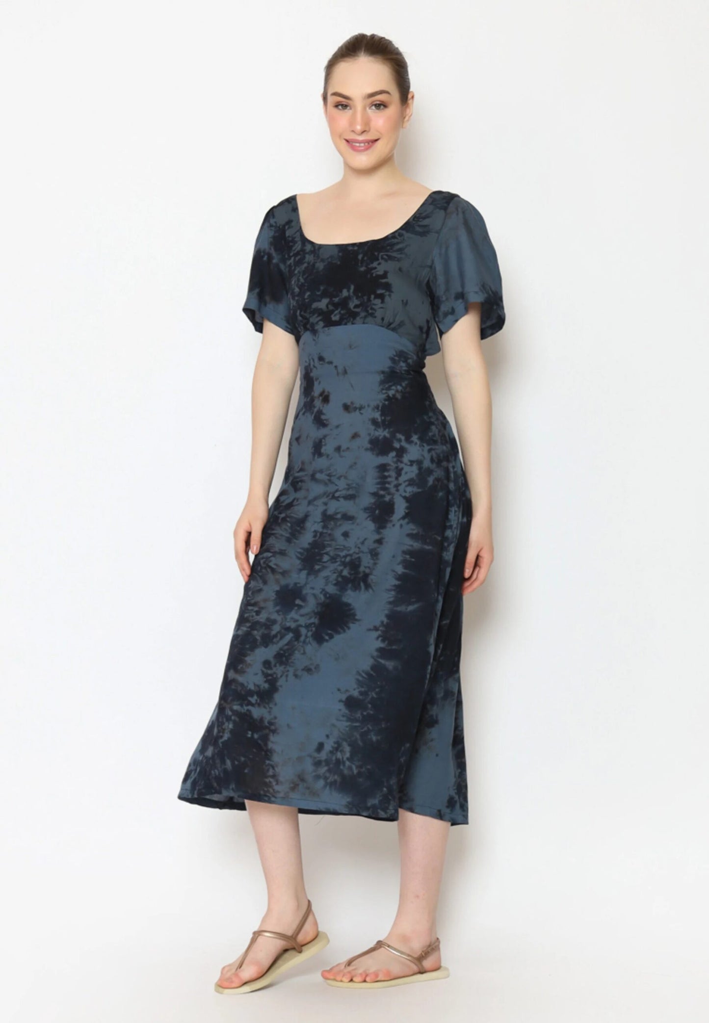 A person smiles, standing against a plain background in a striking tie-back dress. The blue-and-black tie-dye outfit features short sleeves and a square neckline. They complement the look with sandals, and their hair is neatly tied back.