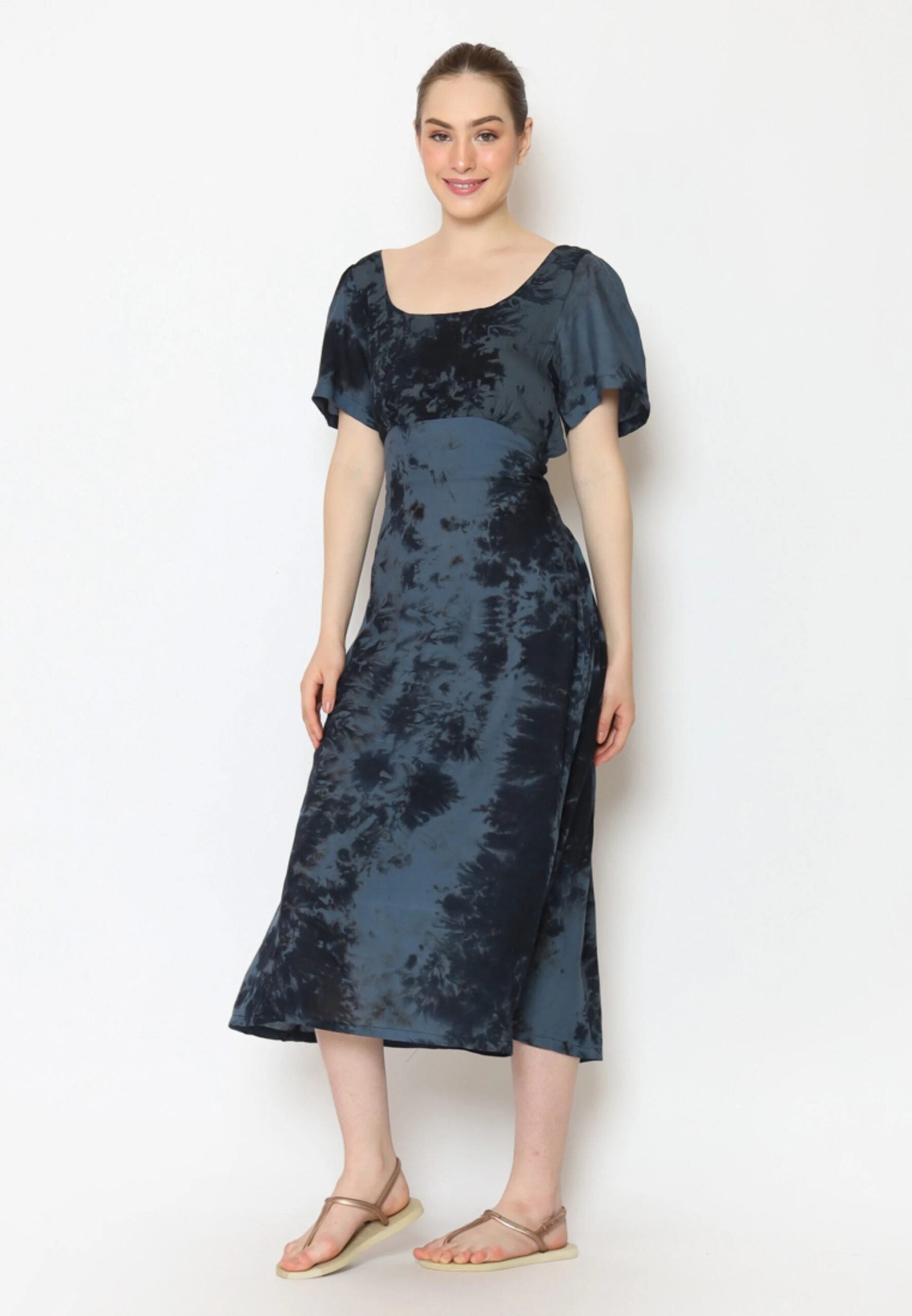 A person smiles, standing against a plain background in a striking tie-back dress. The blue-and-black tie-dye outfit features short sleeves and a square neckline. They complement the look with sandals, and their hair is neatly tied back.