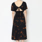 A woman is wearing a black tie back dress adorned with orange floral patterns. The dress features a low, open back with delicate tie details and puffed short sleeves. She stands on a plain white background, her hair in a bun, and complements the look with flat sandals.