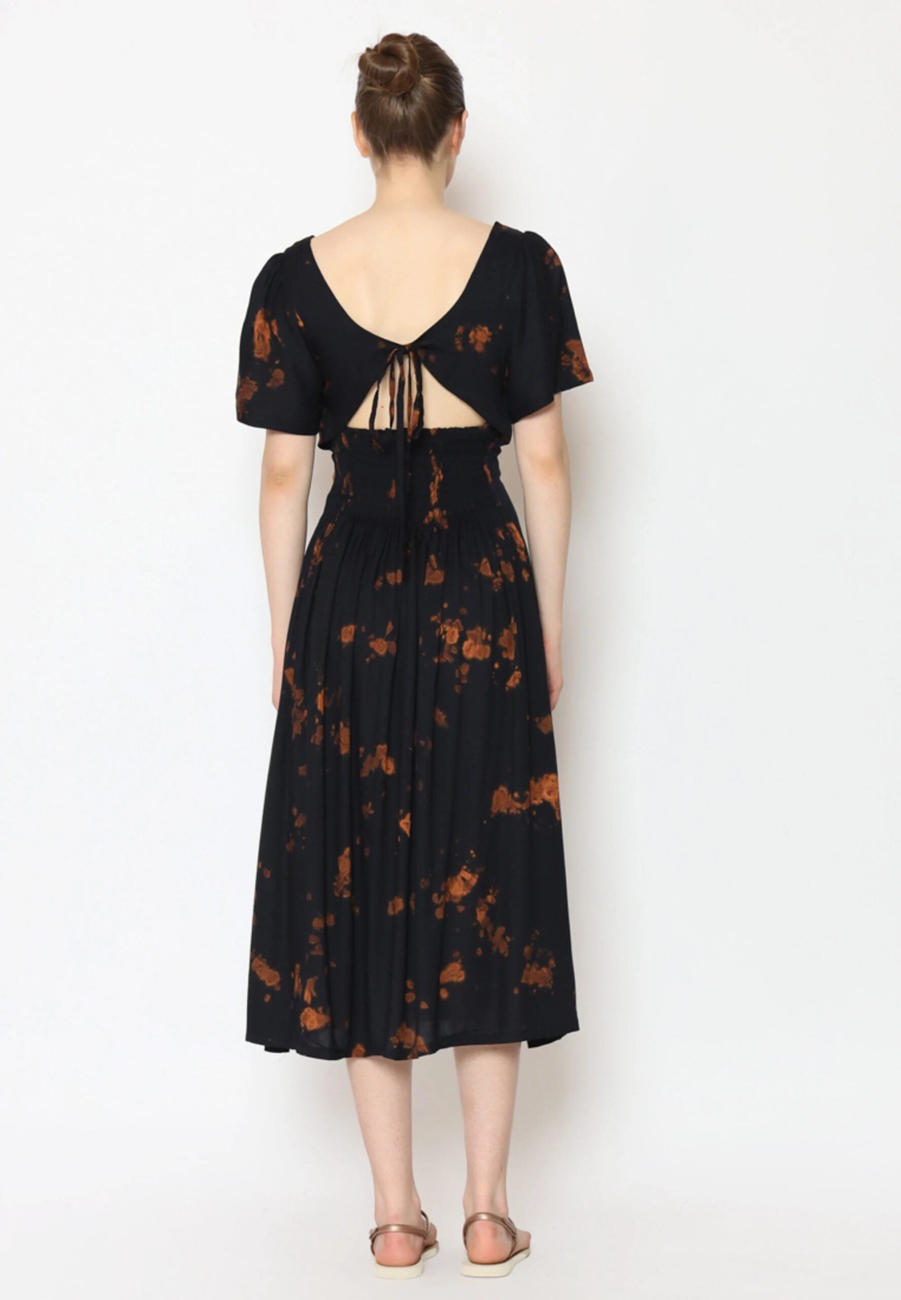 A woman is wearing a black tie back dress adorned with orange floral patterns. The dress features a low, open back with delicate tie details and puffed short sleeves. She stands on a plain white background, her hair in a bun, and complements the look with flat sandals.