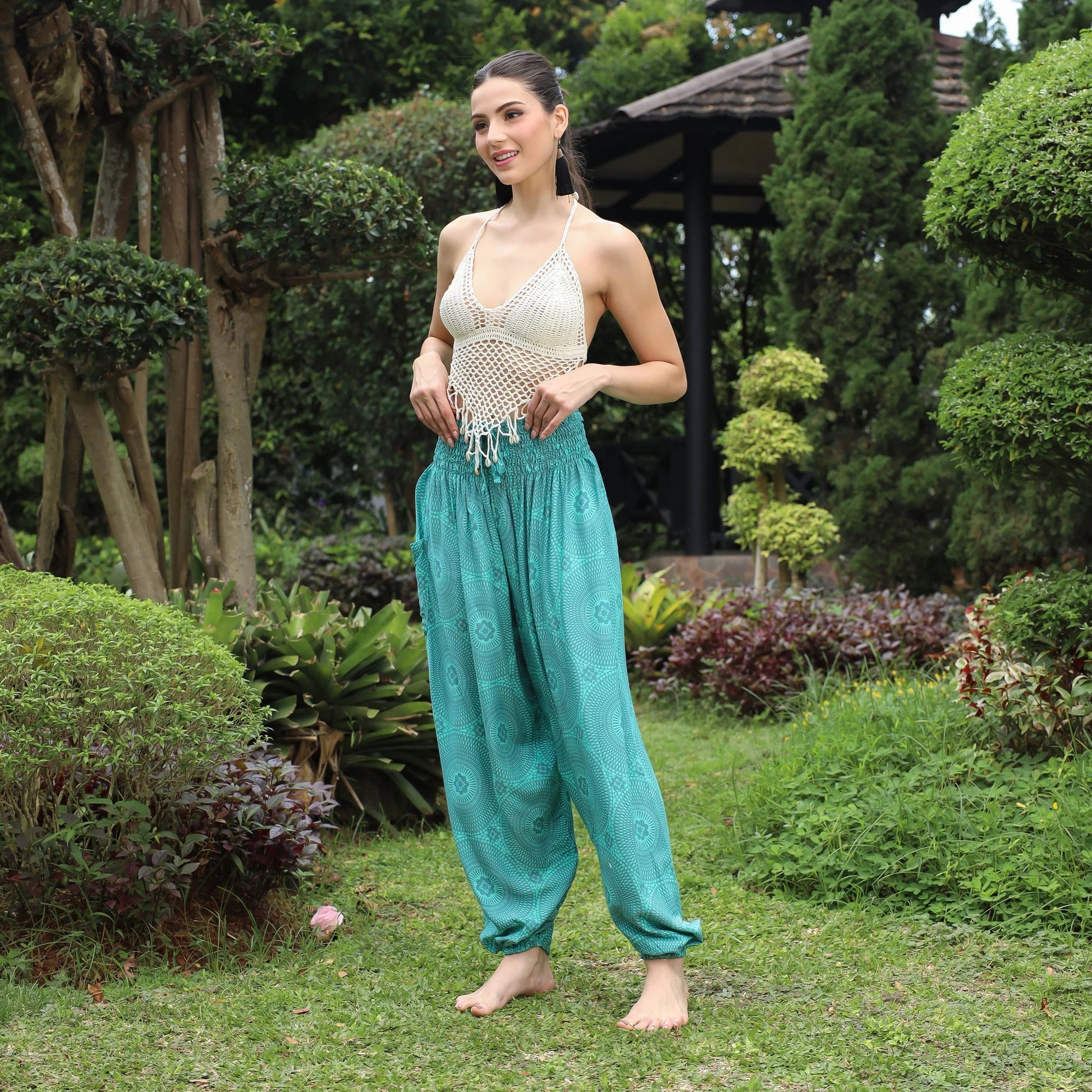 A woman stands barefoot on grass in a garden, wearing a crocheted white top and loose, flowing yoga pants for women. Surrounded by lush greenery and trees, with a gazebo nestled in the background, she exudes serenity.