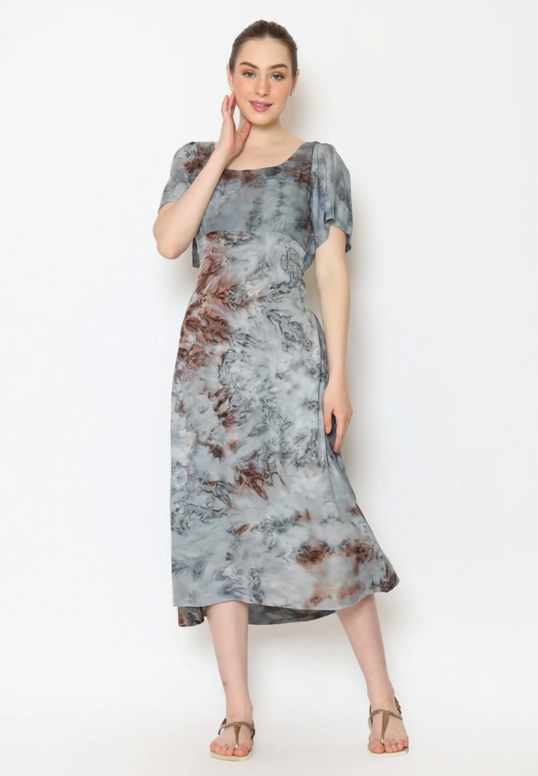 A person stands against a plain background wearing a stylish gray and brown tie back dress. The dress features short sleeves and a mid-length hem. They are smiling, touching their face with one hand, and wearing sandals.