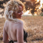 A person with long blonde hair smiles while standing in a sunlit field, wearing a backless crochet top adorned with shells. A tattoo graces their arm as trees and the warm glow of the setting sun paint the background.