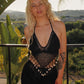 A person with long blonde hair stands outdoors in a black crochet top with shells. They sport a tattoo on their arm, surrounded by lush green plants and scenic hills in the background.