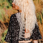 A person with long, wavy blonde hair stands outdoors, wearing a patterned black and white shirt with cut-out details. The background features lush greenery and blurred foliage, suggesting a natural setting.