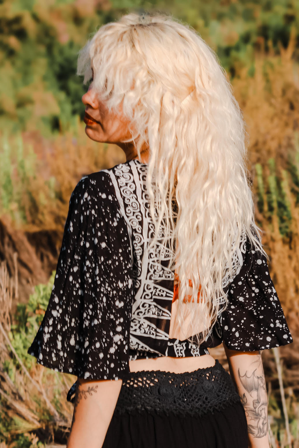 A person with long, wavy blonde hair stands outdoors, wearing a patterned black and white shirt with cut-out details. The background features lush greenery and blurred foliage, suggesting a natural setting.