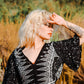 A person with curly blonde hair and tattoos on their arm stands outdoors, shielding their eyes from the summer sun with one hand. They are wearing a black and white patterned top, perfect for summer tops for women, surrounded by dry grass and blurred greenery in the background.