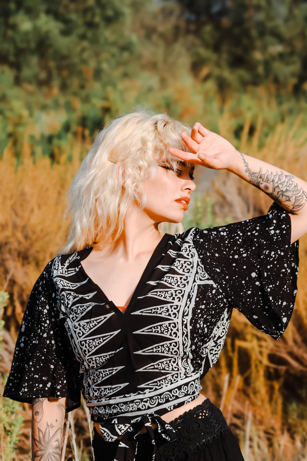 A person with curly blonde hair and tattoos on their arm stands outdoors, shielding their eyes from the summer sun with one hand. They are wearing a black and white patterned top, perfect for summer tops for women, surrounded by dry grass and blurred greenery in the background.