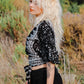 A woman with long, wavy blonde hair stands in a field of tall grass, wearing one of the latest summer tops for women—a stylish black and white patterned number—paired with black pants. The background is filled with lush greenery under a clear sky.
