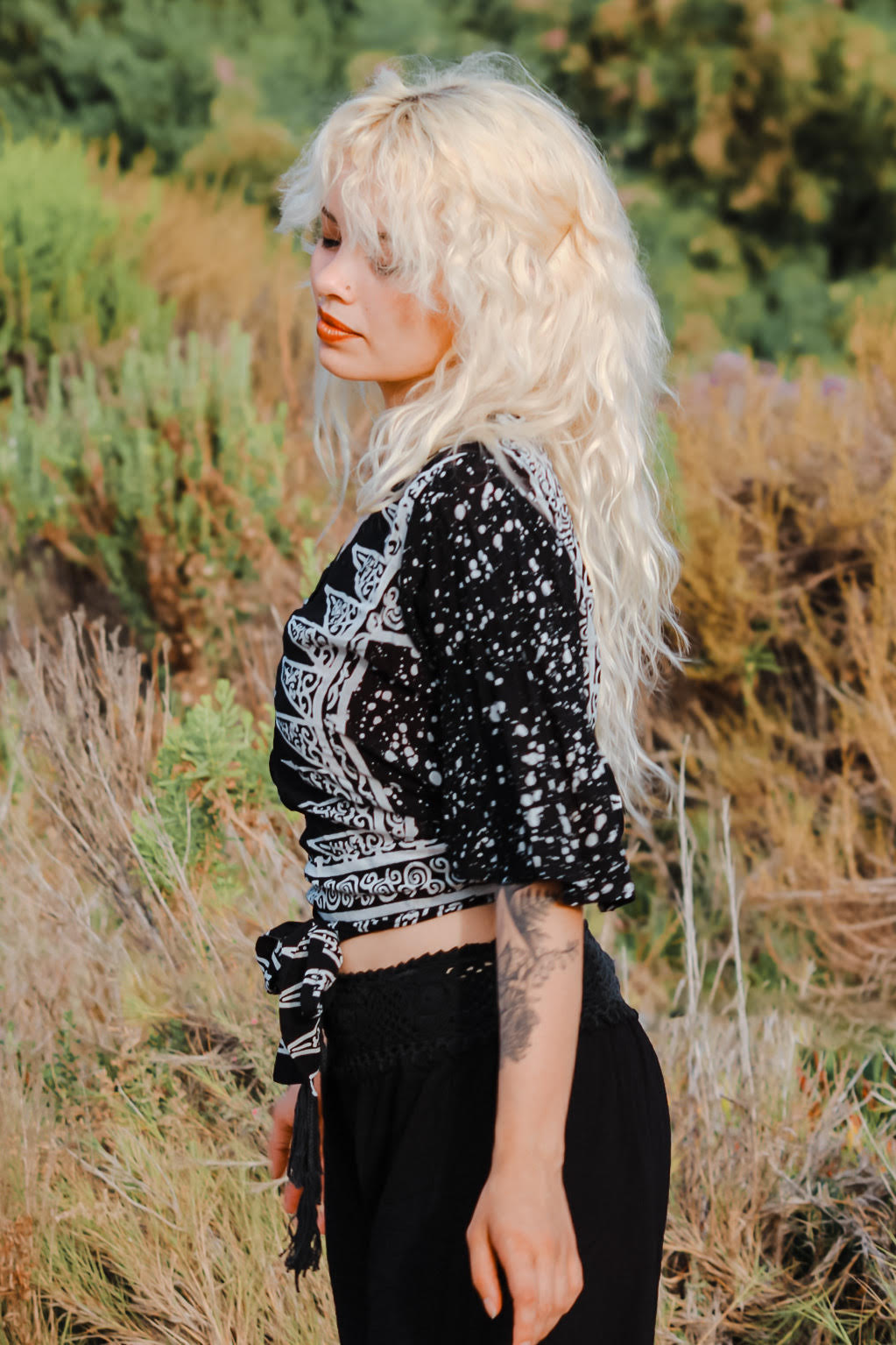 A woman with long, wavy blonde hair stands in a field of tall grass, wearing one of the latest summer tops for women—a stylish black and white patterned number—paired with black pants. The background is filled with lush greenery under a clear sky.